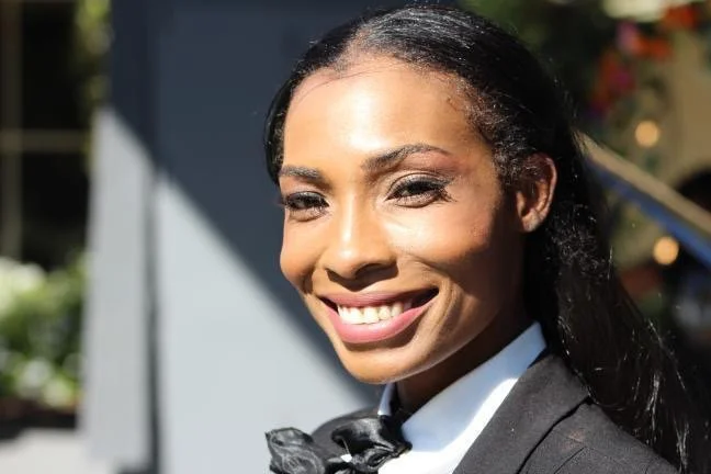 A smiling woman with dark hair styled straight, wearing a black blazer and a white shirt with a black bow tie, outdoors on a sunny day.