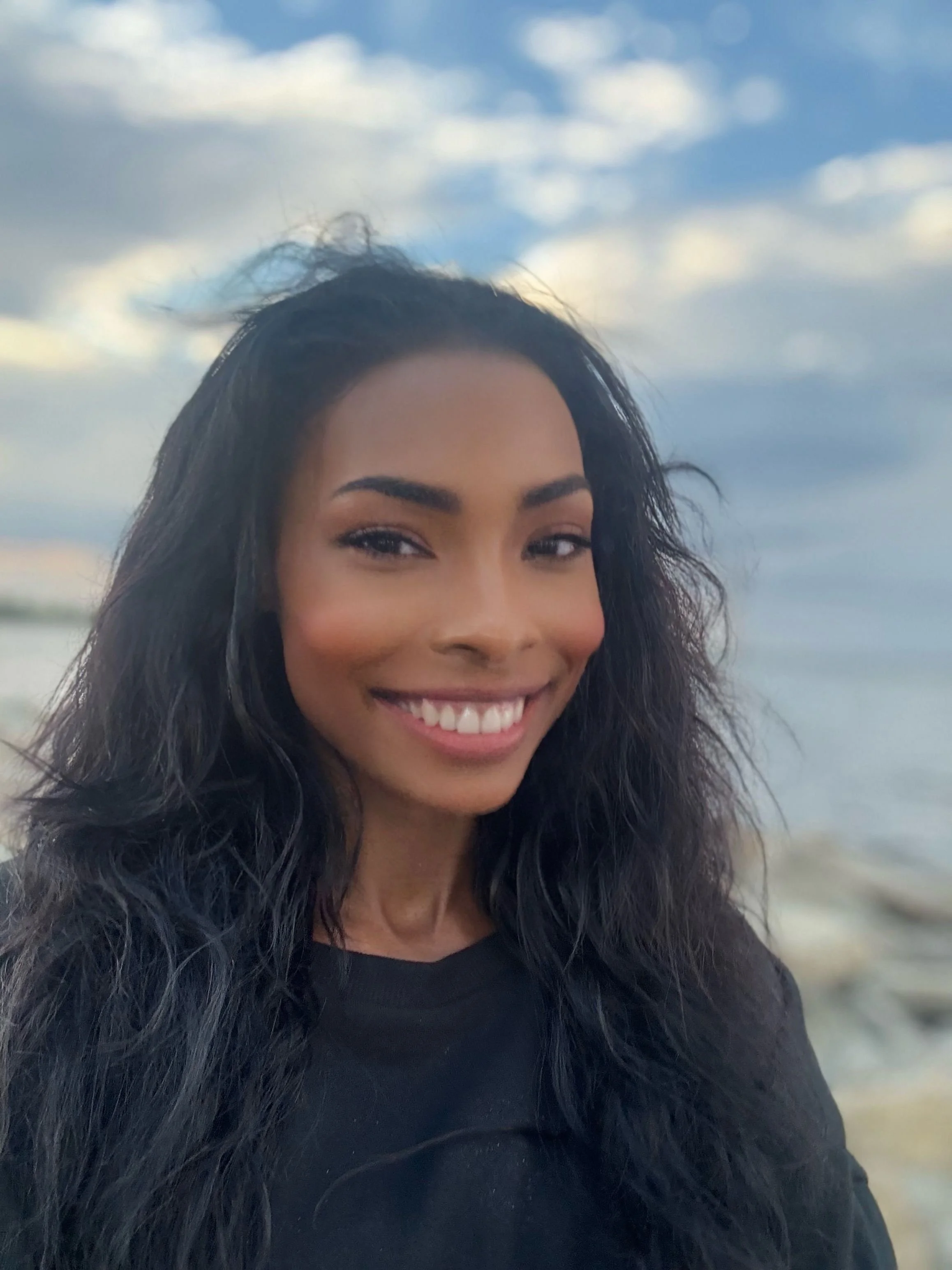 A woman with dark curly hair smiling at the camera, with a cloudy sky and a body of water in the background.