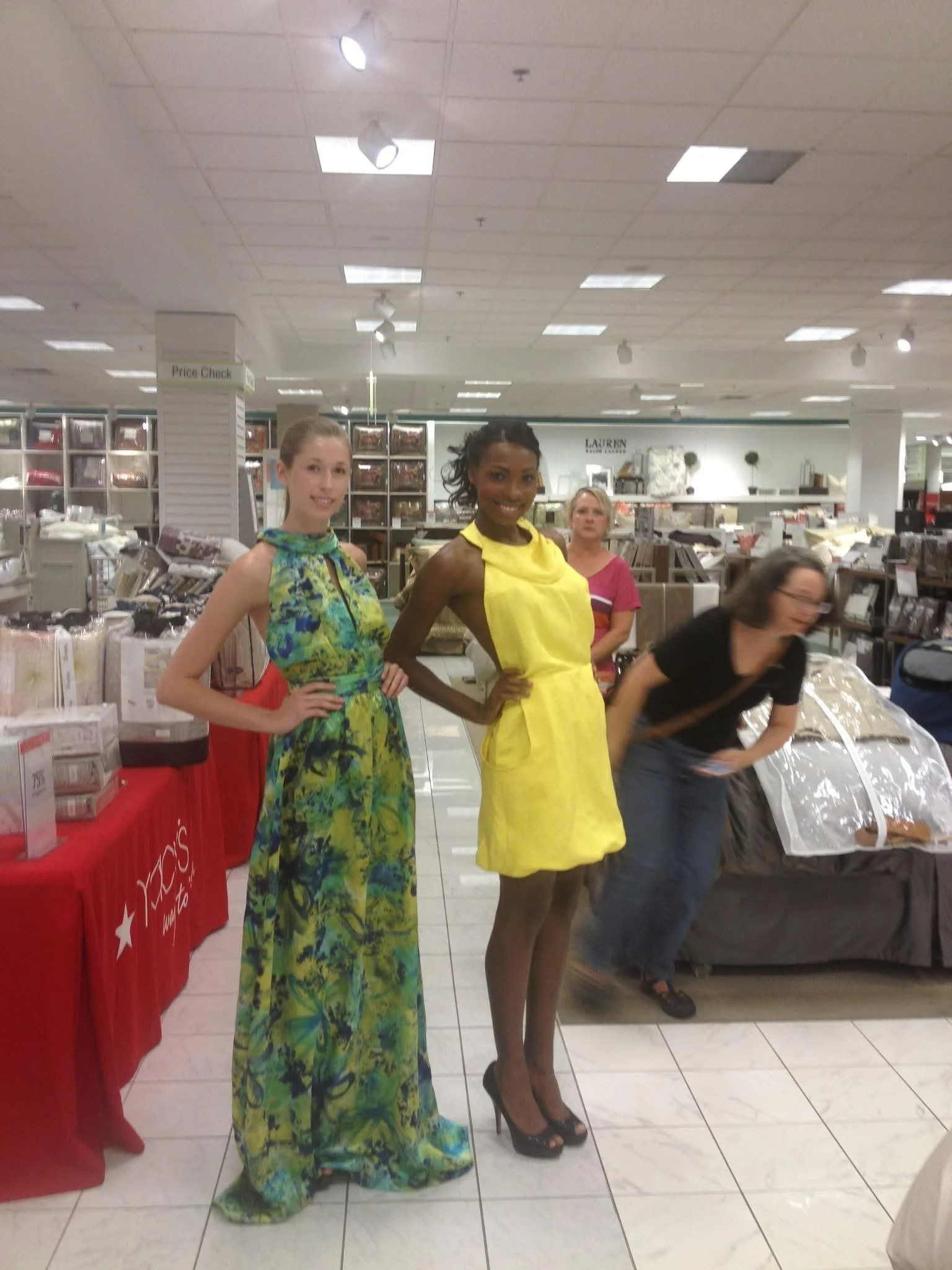 Four women standing inside a store, with two women in the foreground wearing colorful dresses and high heels, and two women in the background near shelves of household items and a checkout area.