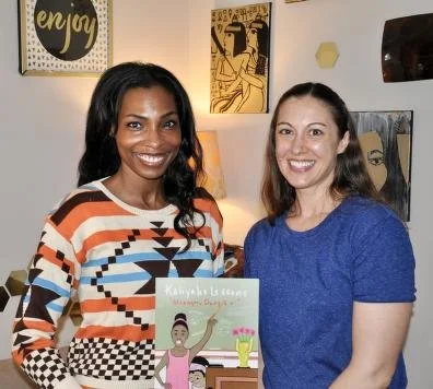 Two women standing indoors, smiling, with artwork on the wall behind them, one holding a colorful book.