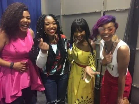 Four women smiling and laughing together in a room with a gray wall.