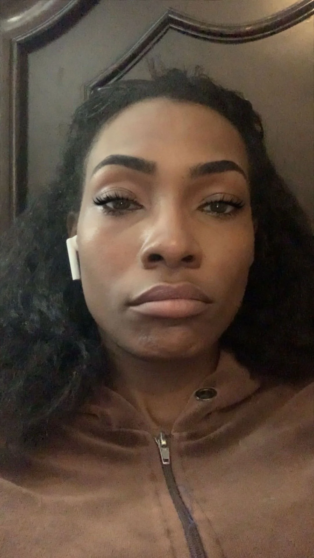 Close-up of a woman with dark, curly hair, wearing makeup, a brown jacket with a zipper, and white wireless earbuds, lying on a headboard or bed.