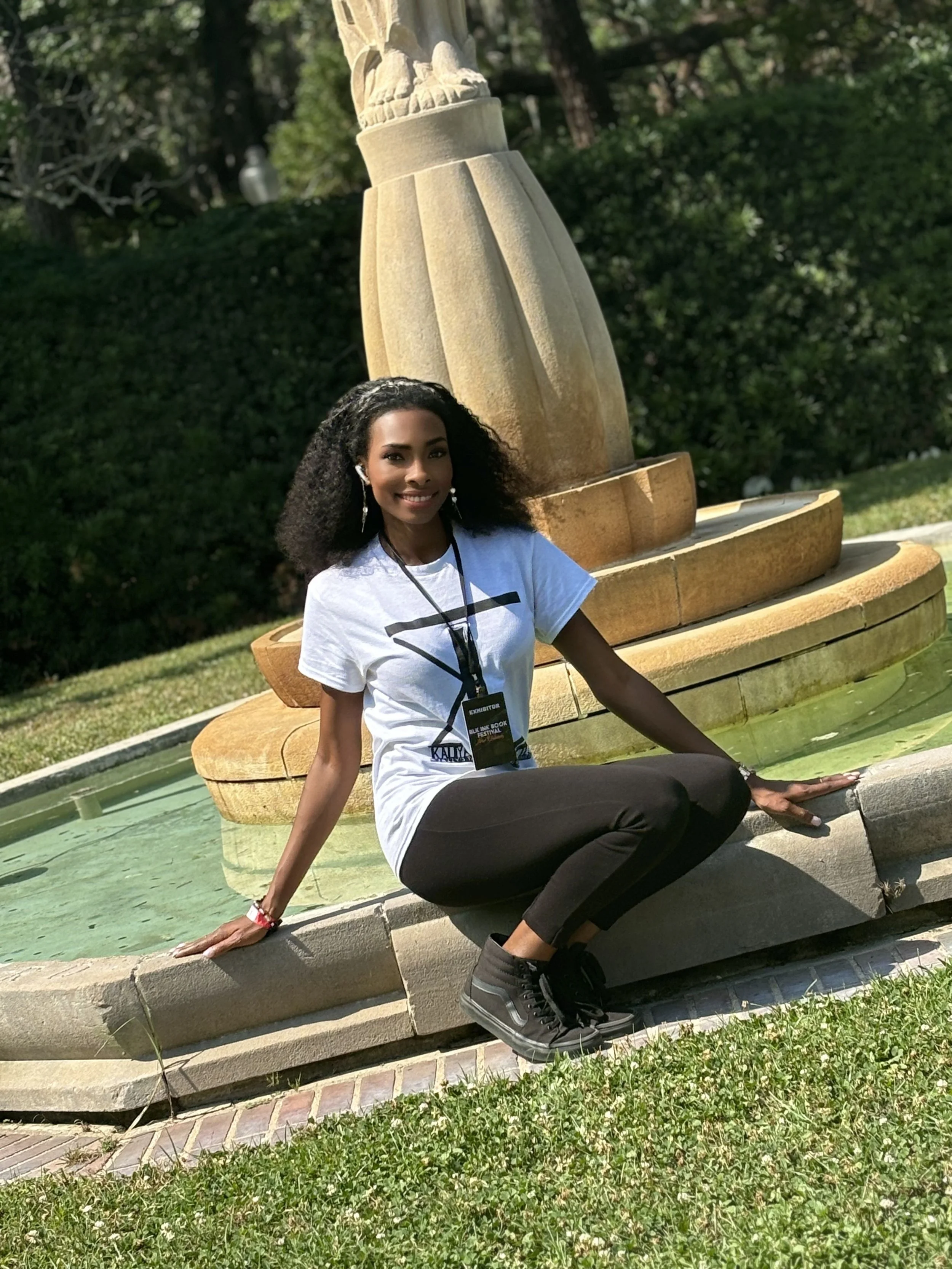 A young woman with curly hair, wearing a white t-shirt, black pants, and black sneakers, is sitting on a rounded stone edge of a fountain in a park, smiling at the camera. The fountain has a decorative sculpture on top and is surrounded by greenery.