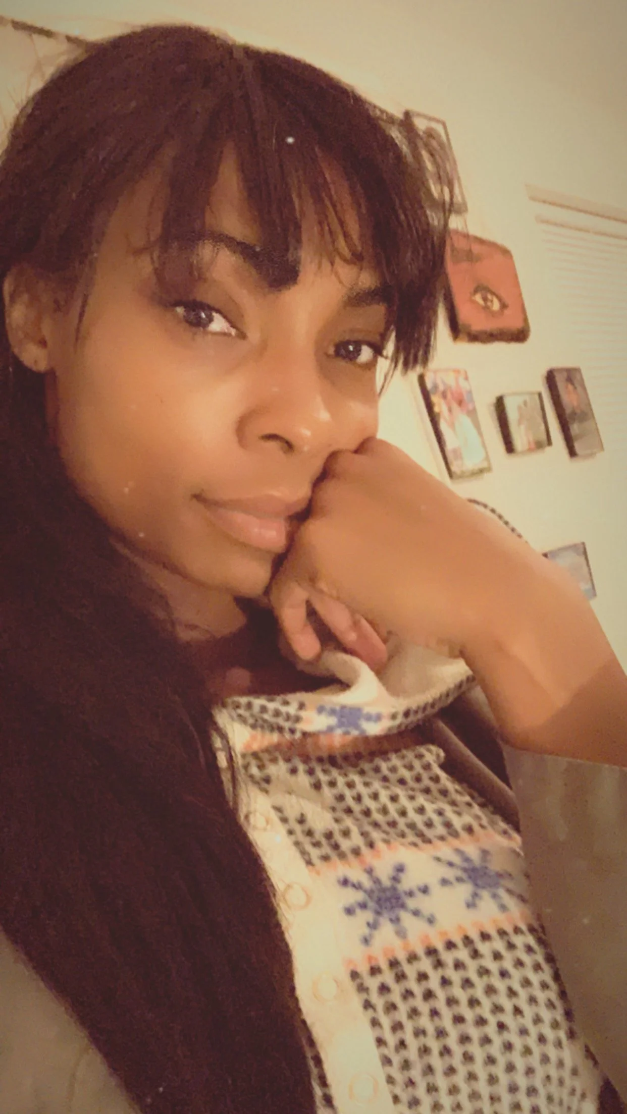 A woman with dark hair and makeup, resting her chin on her arm, sitting in a room decorated with framed pictures and artwork on the wall.