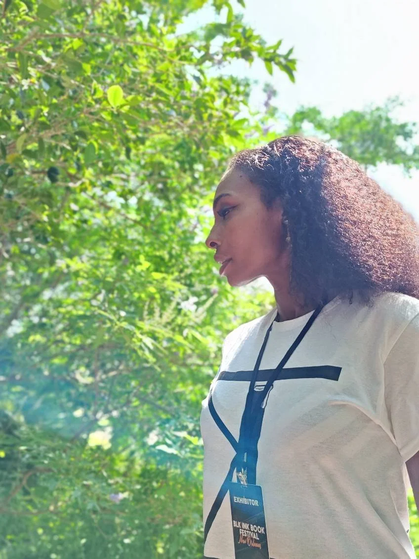 Profile of a woman with curly hair wearing a white T-shirt and a lanyard with an exhibitor badge, standing outdoors with green foliage and sunlight in the background.