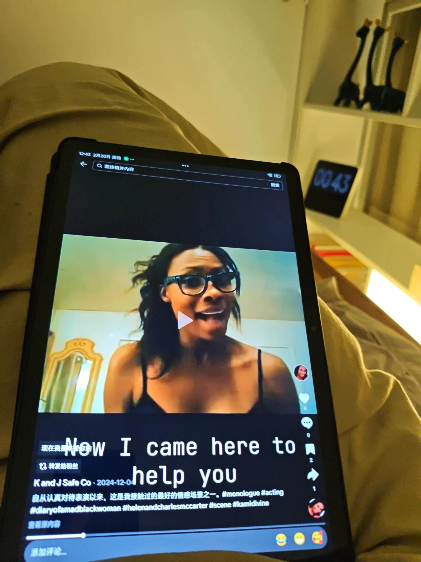 A person holding a tablet, displaying a video or photo of a woman with glasses and black clothing. In the background, there is a digital clock showing 00:45 and a shelf with three giraffe figurines.