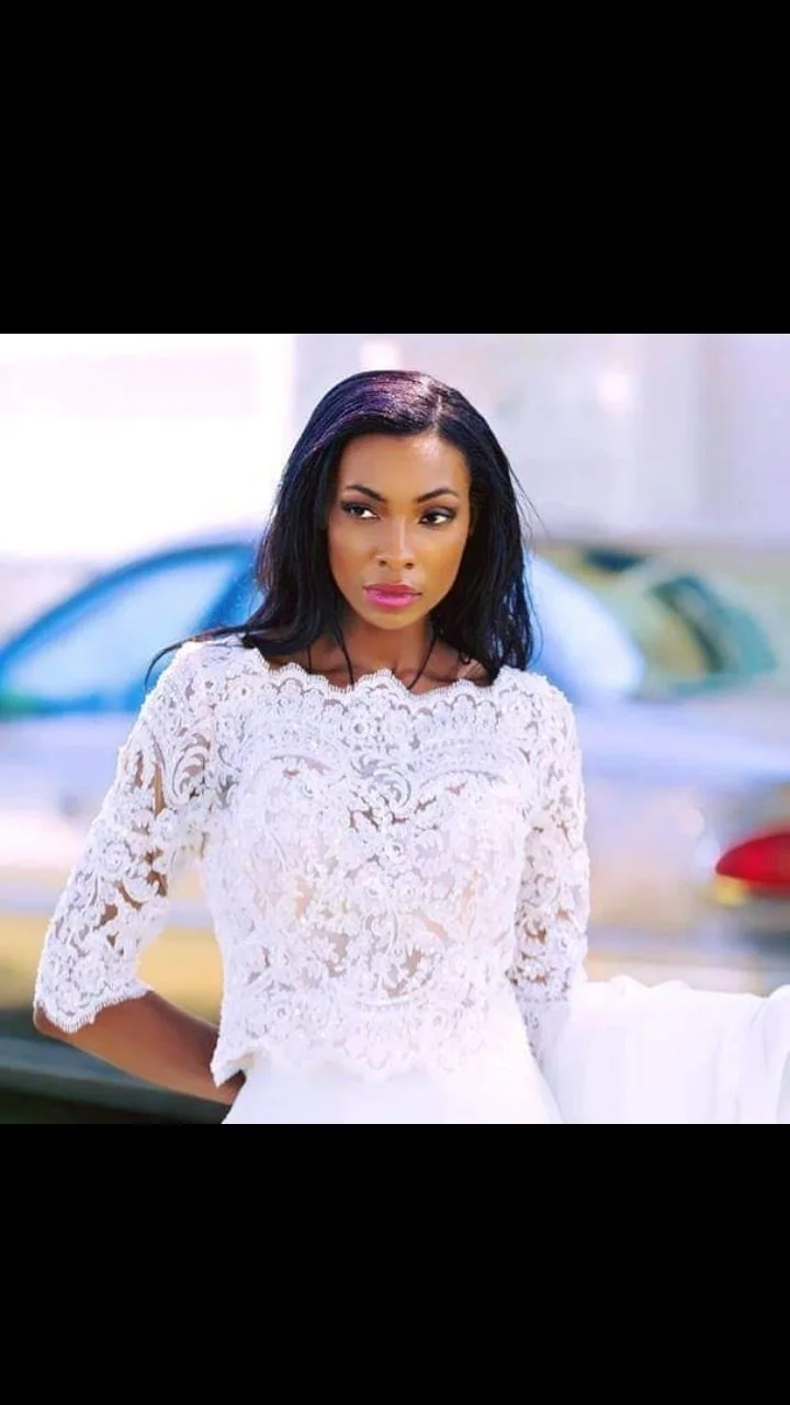 A woman with dark hair and pink lipstick wearing a white lace dress outside with blurred cars in the background.