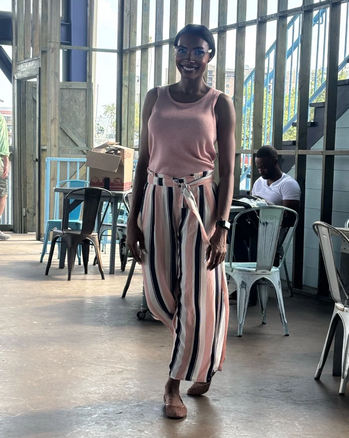 A woman walking inside a modern, industrial-style cafe with metal chairs and wooden walls. She is smiling, wearing glasses, a pink sleeveless top, and high-waisted, wide-leg striped pants tied at the waist. There is a man sitting in the background wo
