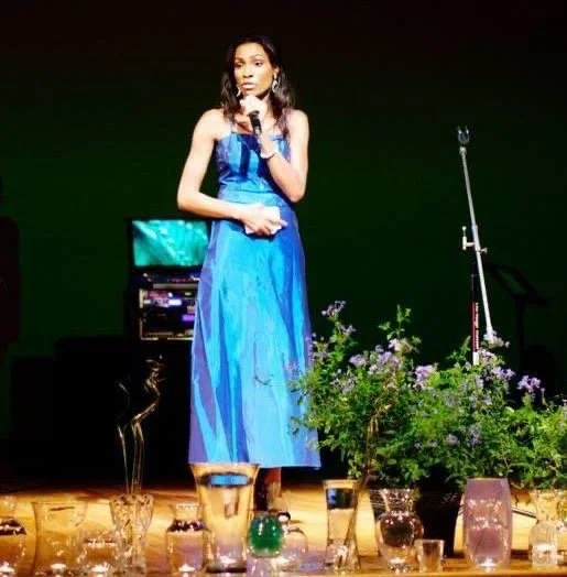 A woman in a long blue dress standing on a stage, holding a microphone, surrounded by flower arrangements and glassware.