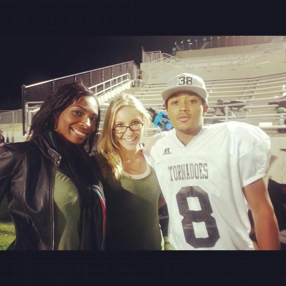 Three people, two women and a young man in football uniform, standing together on football field at night.