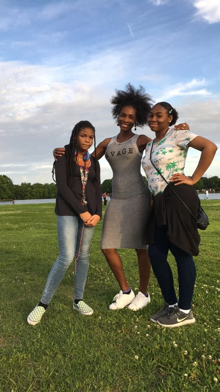 Three women standing on a grassy field near water, smiling and posing for a photo. The woman in the middle is wearing a gray dress, the woman on the right is wearing a tropical print shirt, and the woman on the left has long braided hair and is dress
