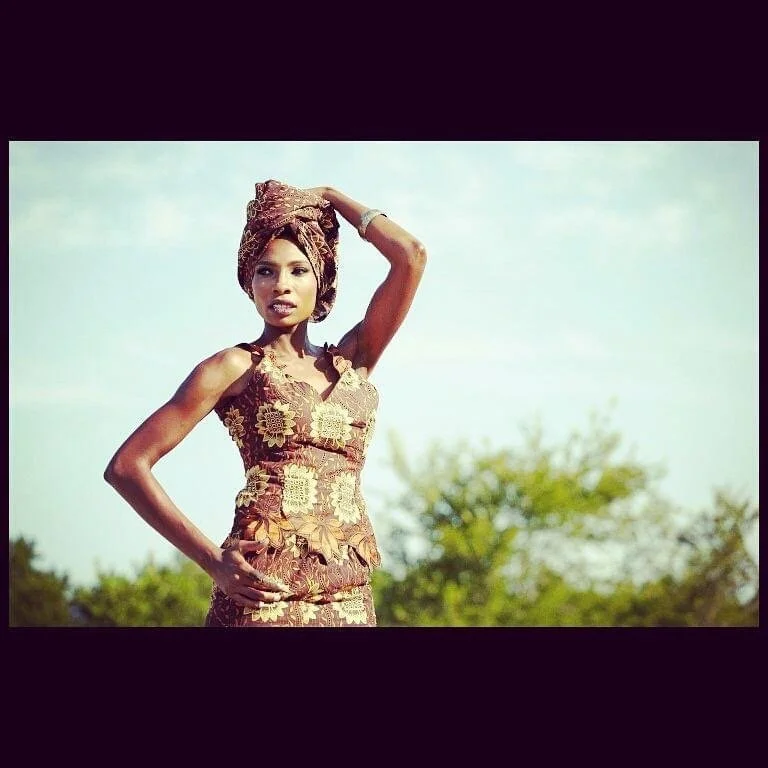 A woman dressed in traditional African attire with a patterned headwrap, standing outdoors with a blurred natural background.