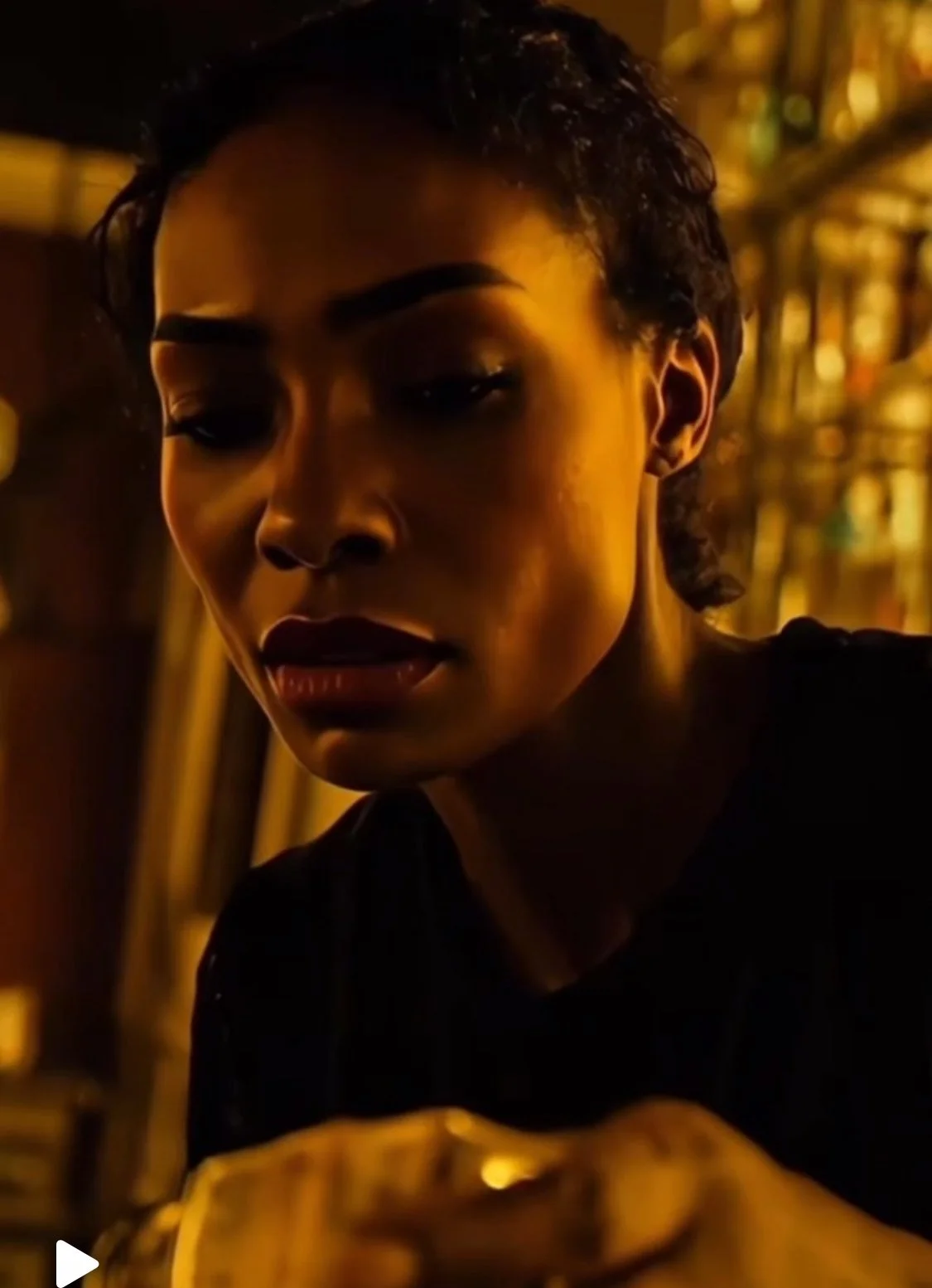 A woman with short dark hair looking downward, illuminated by warm yellow lighting, with a blurry background of shelves and bottles.