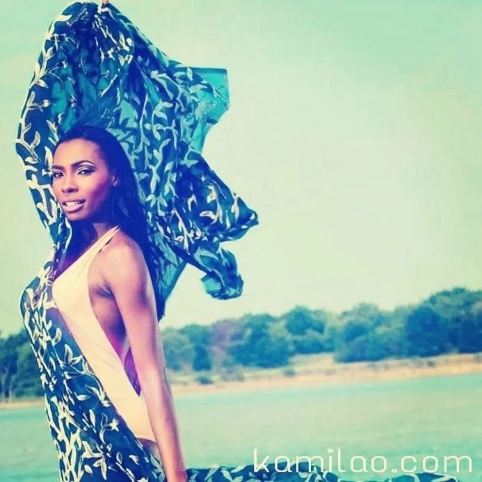 A woman standing outdoors near water, holding a large patterned fabric or scarf over her head, wearing a yellow swimsuit.