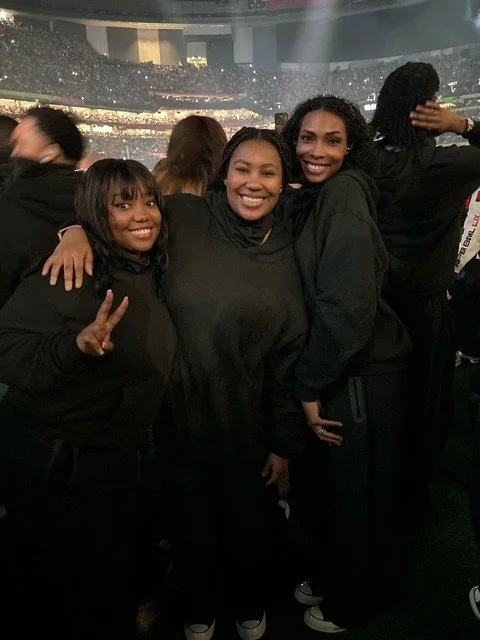 Three women smiling and posing for a photo at a concert or event, with a crowd and stage in the background.