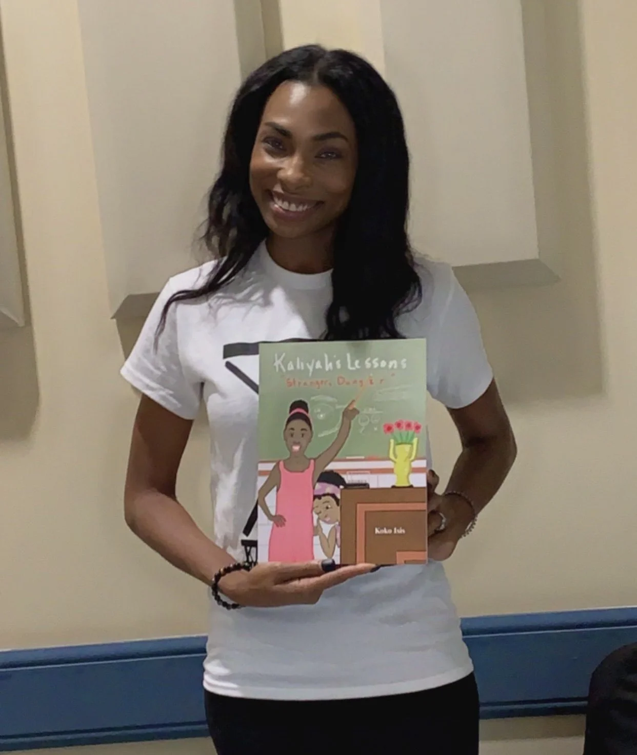 A smiling woman with dark hair holding a children's book titled 'Kaliyah's Lessons: Struggles, Dagger'. She is wearing a white T-shirt and standing indoors against a cream-colored wall.