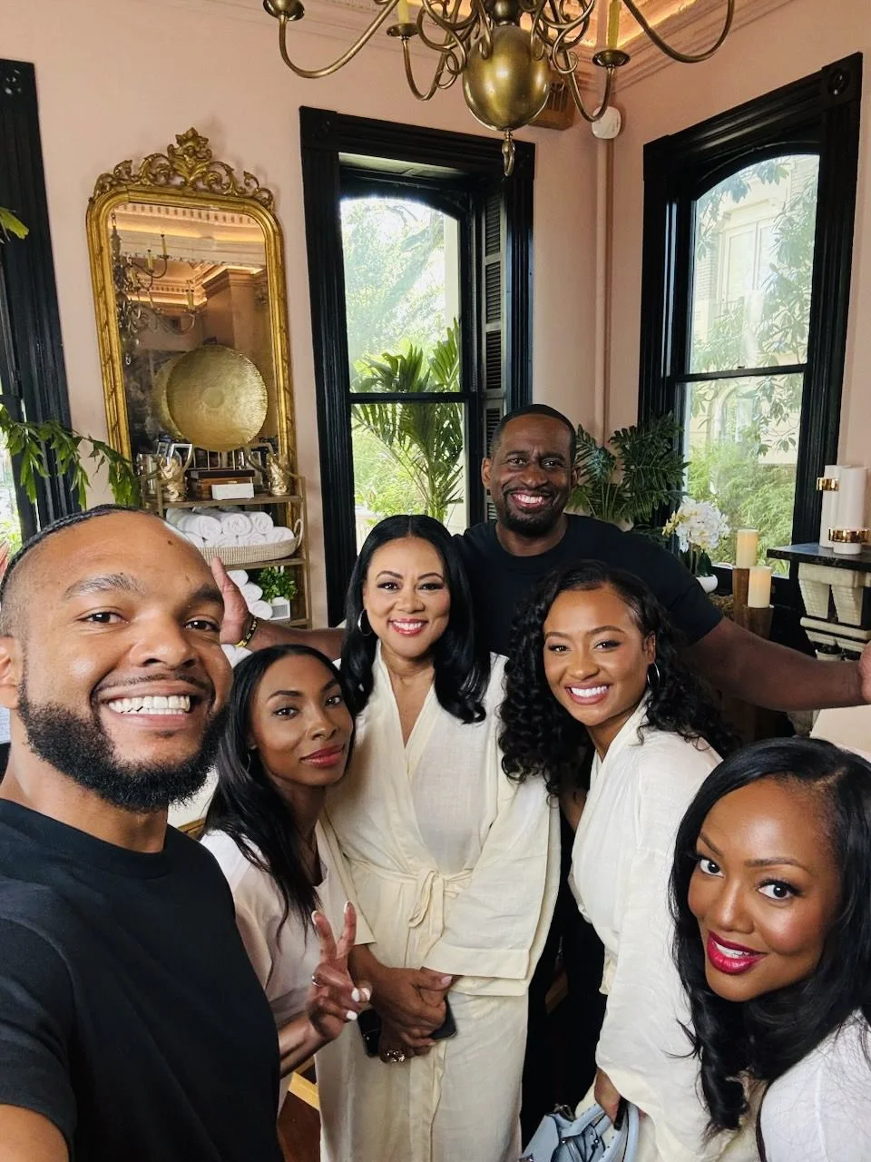 Group of six people smiling and taking a selfie in a well-decorated room with large windows, plants, candles, and a gold mirror.