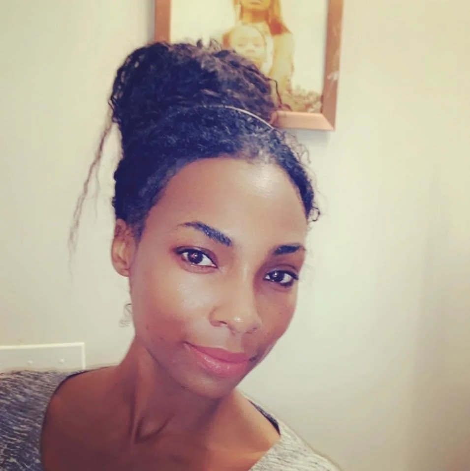 Portrait of a woman with braided hair styled in a high bun, smiling softly, in a room with a framed picture on the wall behind her.
