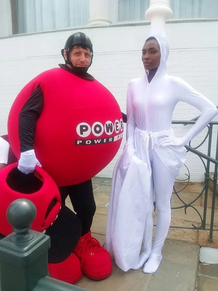 Two people in costumes, one dressed as a large red Powerball lottery ball and the other as a white lamp or genie, posing outdoors near a white building.