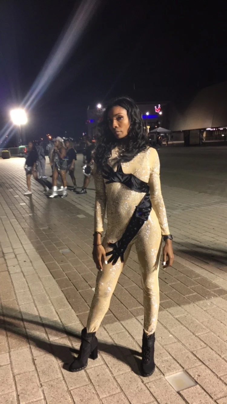 A woman with long curly black hair in a gold and black bodysuit and high-heeled boots, standing outdoors at night on a paved area with people in the background.
