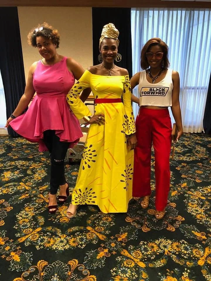 Three women standing on a floral carpeted floor in front of dark and white curtains at an indoor event.