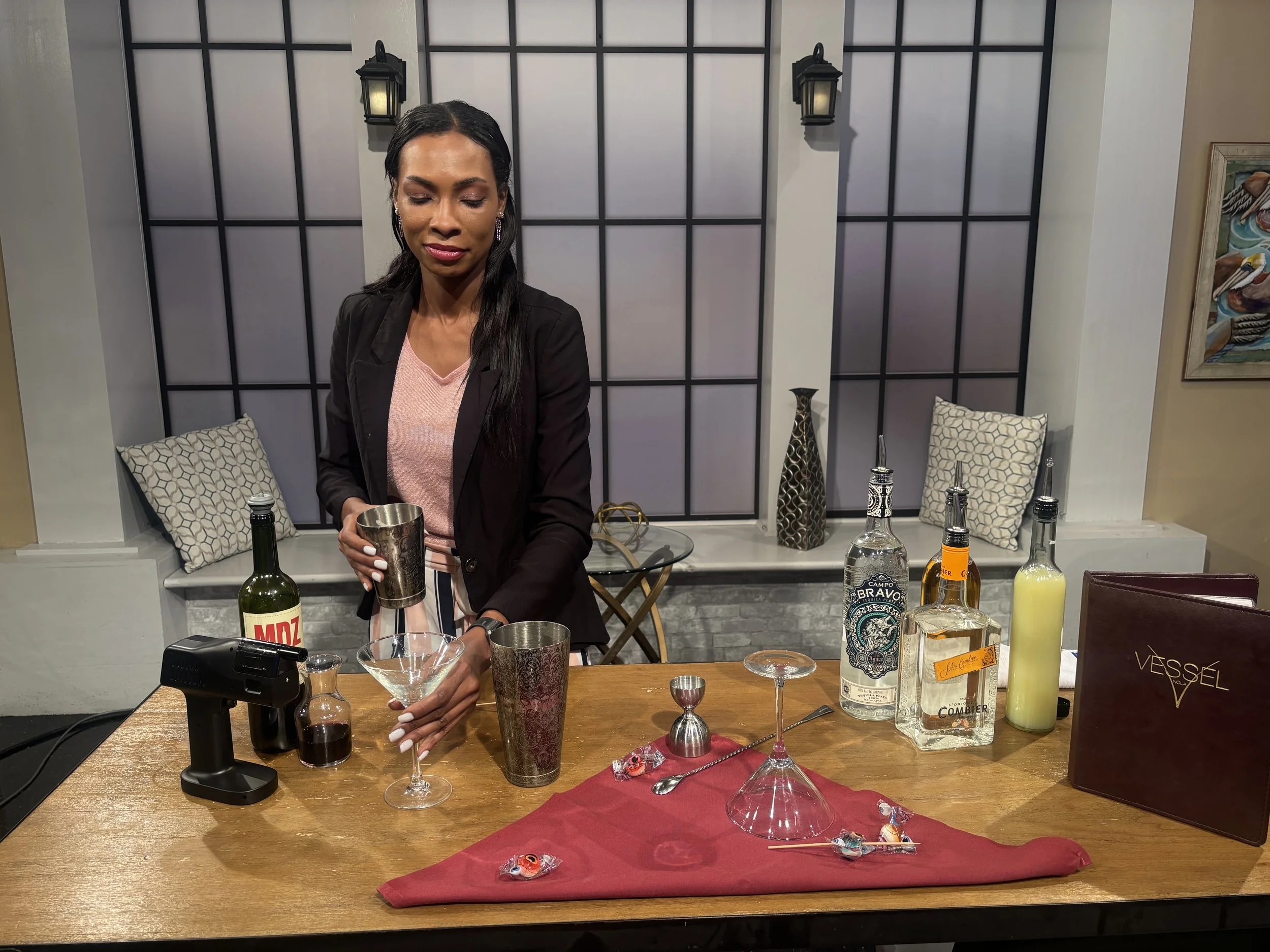 A woman with long dark hair and earrings standing behind a wooden table with various bottles of alcohol, glassware, and candies, in a room with a modern black and white wall, pillows, and artwork on the wall.