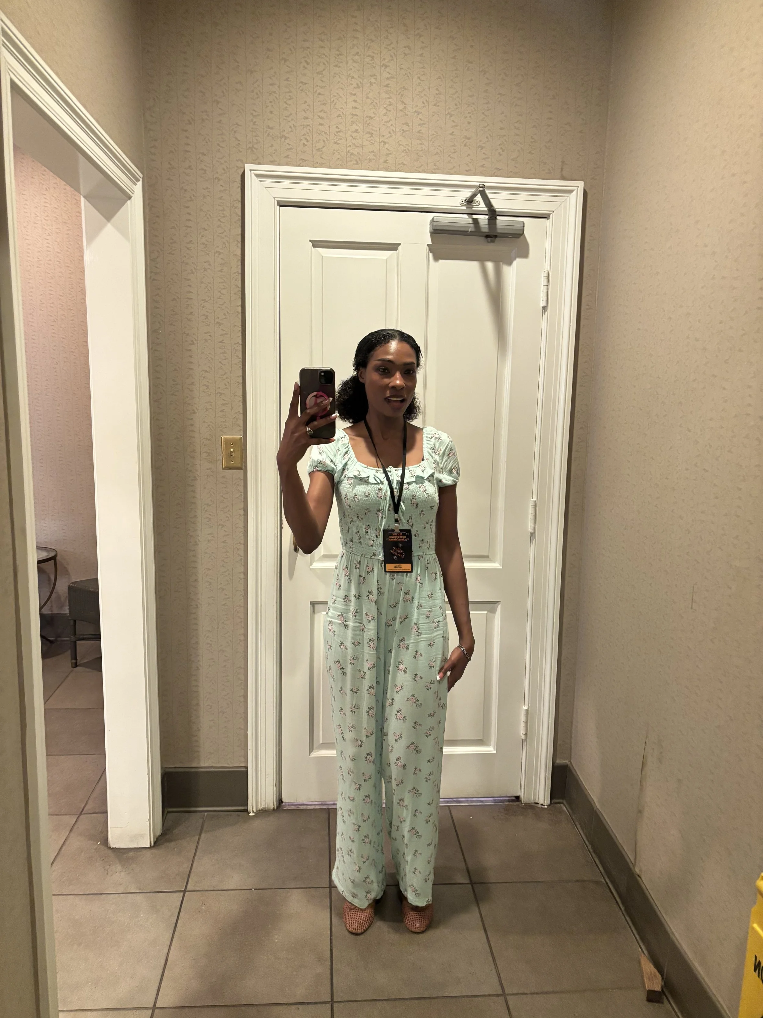 A woman taking a mirror selfie in a fitting room, wearing a light green floral jumpsuit and pink shoes.