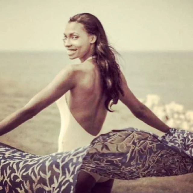 A woman with long brown hair sitting on a beach, smiling, with an ocean backdrop and a cloudy sky.