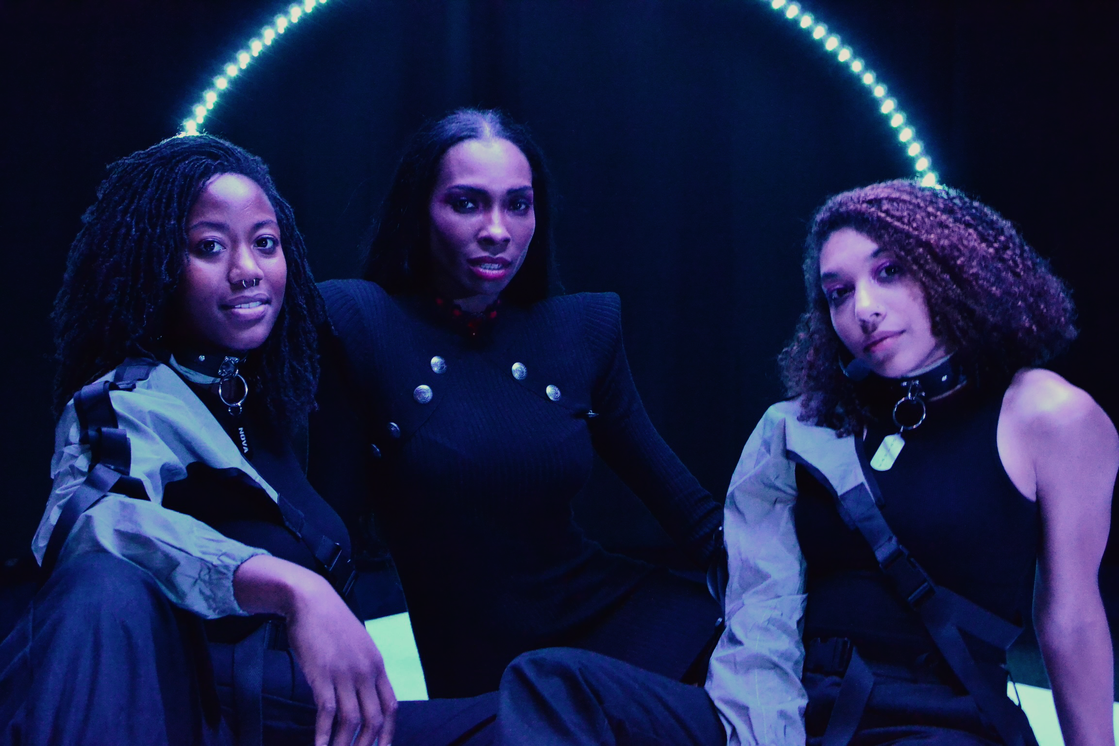 Three women dressed in dark and light jackets posing together in a dark room with blue and purple lighting.