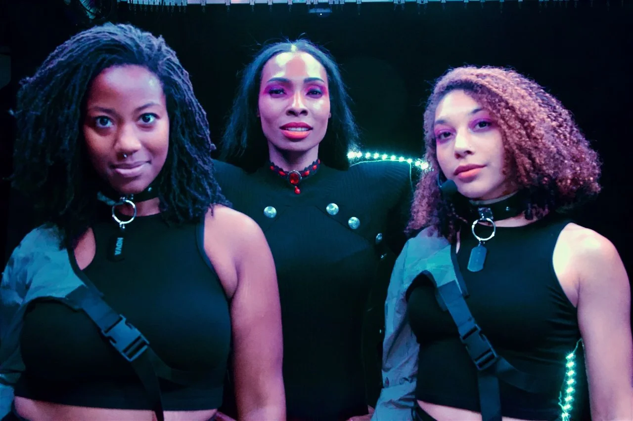 Three women with distinctive hair styles posing together in dark environment with vibrant lighting.