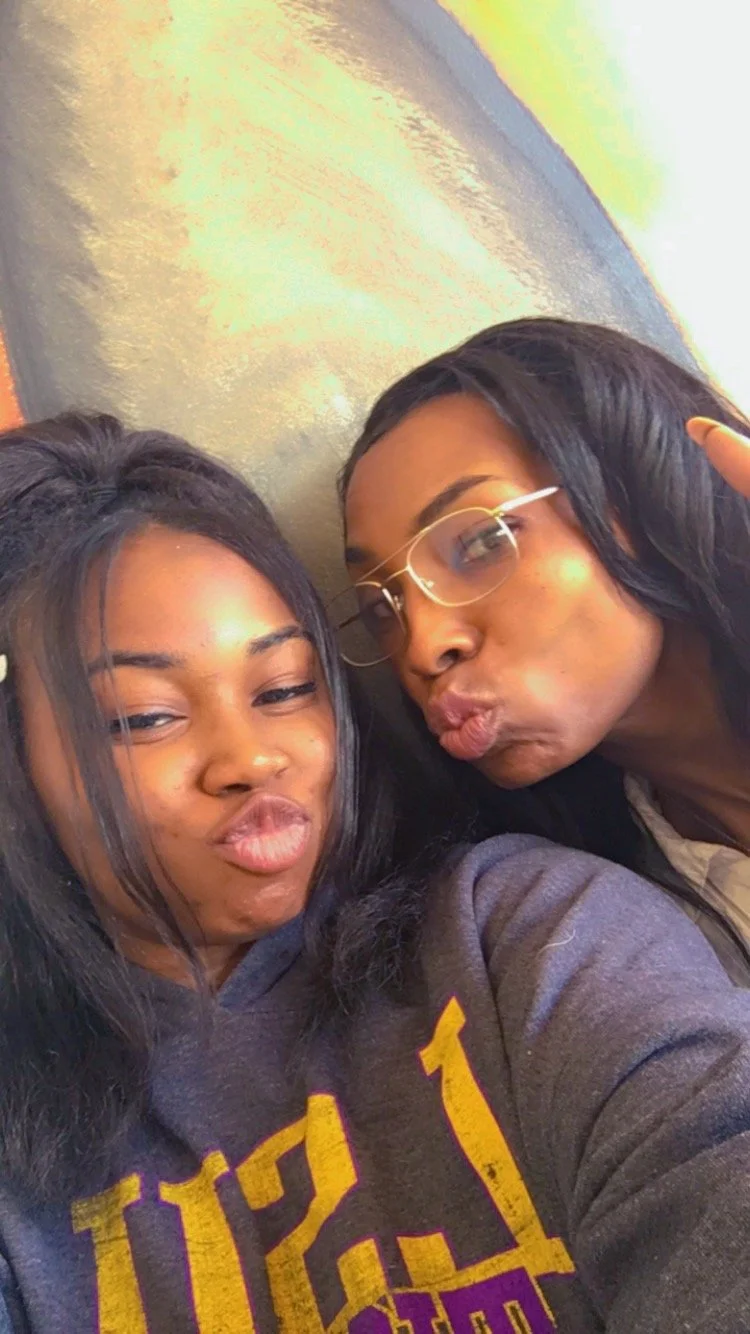Two young women taking a selfie together, making funny face expressions, against a textured wall background.