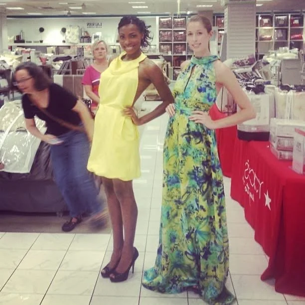 Two women standing side by side in a store, one in a yellow dress and the other in a long multicolored dress, with two other women in the background near a display table.