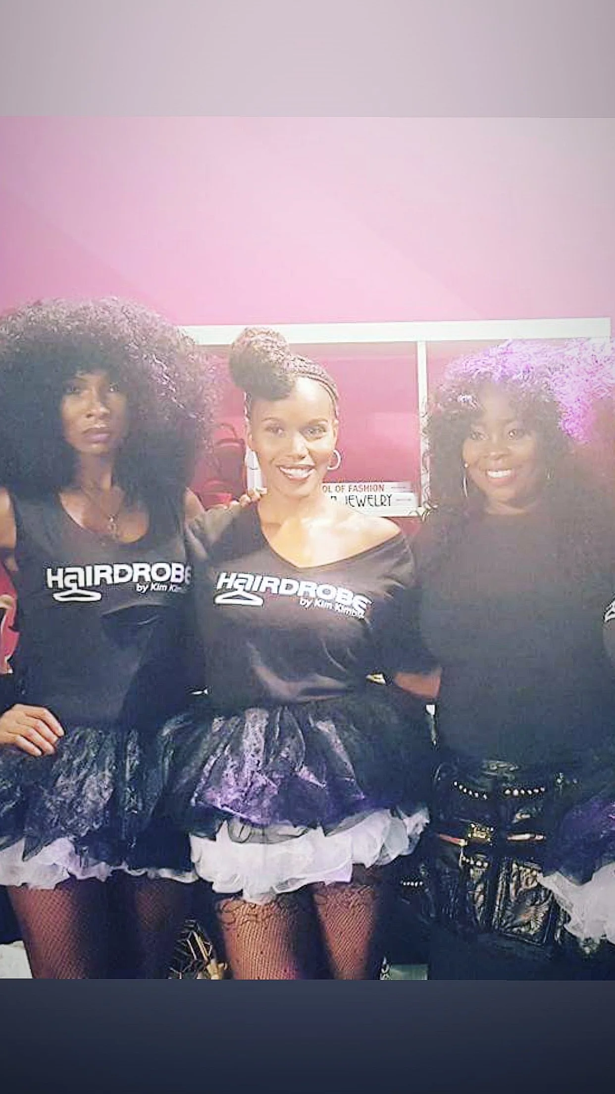Three women standing together in fashionable outfits. They are in front of a pink wall and a sign that says "School of Fashion and Jewelry." The women are wearing black T-shirts with "Hair Drop" written on them, and they have unique hairstyles and ac