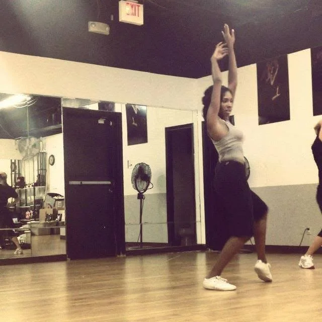 A woman dancing in a studio, with her arms raised above her head and smiling, wearing a gray tank top and black shorts.