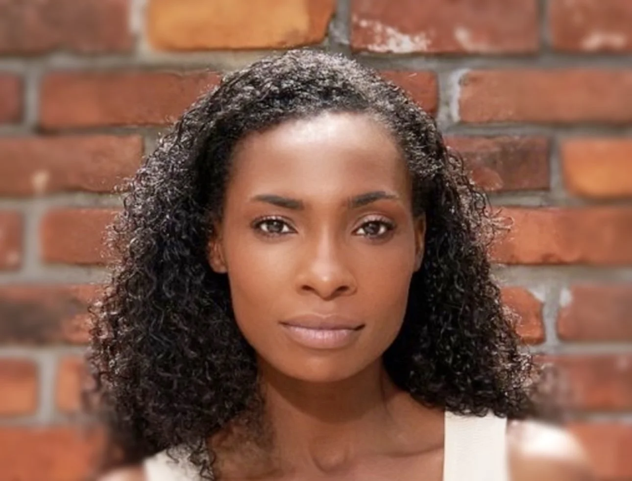 Close-up portrait of a woman with dark curly hair against a brick wall background.
