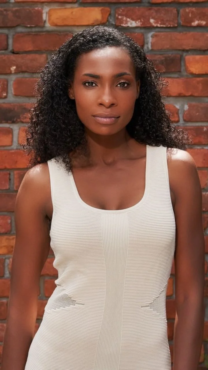 A woman with curly black hair and medium skin tone wearing a sleeveless white textured dress with a brick wall background.