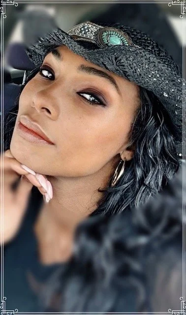 Close-up of a woman wearing a black cowboy hat with turquoise decoration, hoop earrings, and makeup, in a car.