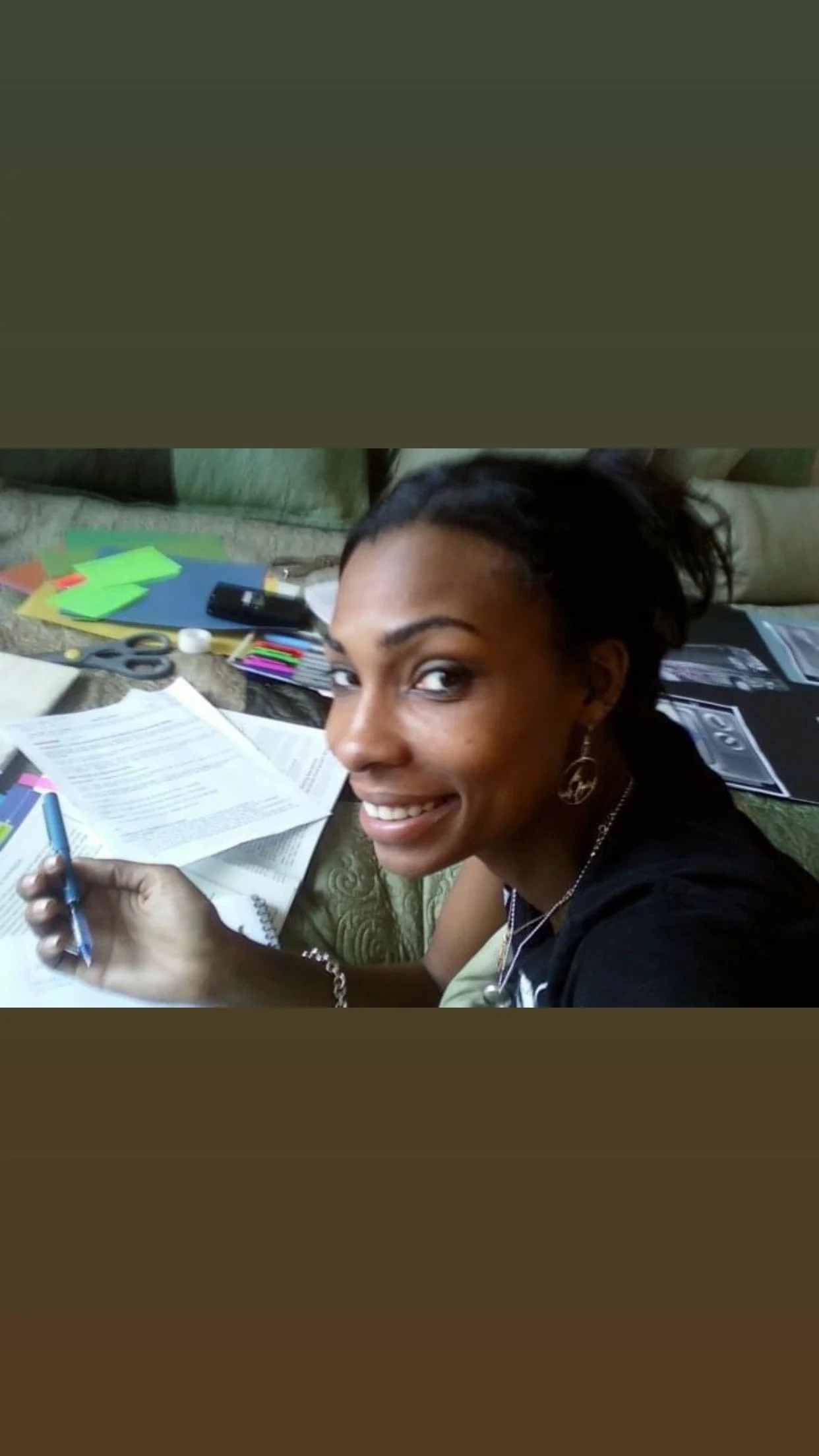A woman with dark hair tied back, smiling at the camera while holding a pen over papers and documents, with colorful sticky notes, scissors, and office supplies on the table in the background.