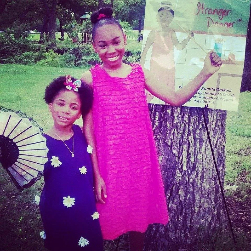 Two young girls standing outdoors with trees in the background. One girl is dressed in a purple dress decorated with white flowers and has a colorful bow in her hair, while the other girl wears a pink dress. The taller girl is smiling and holding a s
