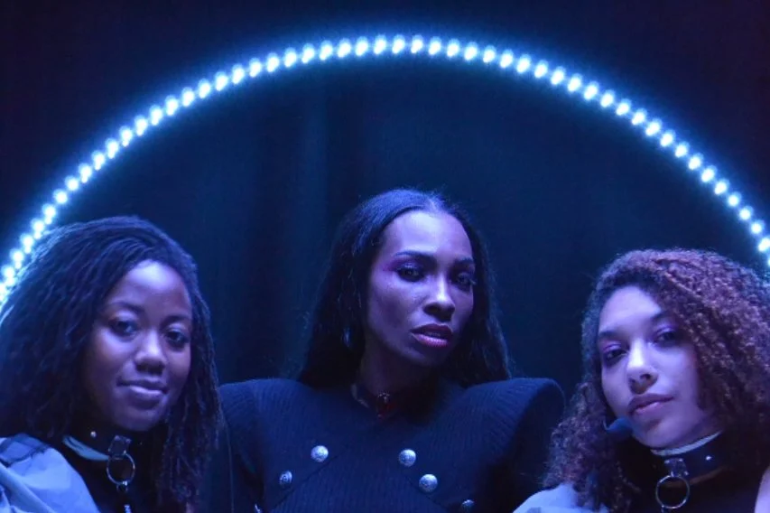 Three women with dark hair and makeup, standing in front of a dark background with blue lights forming an arch.