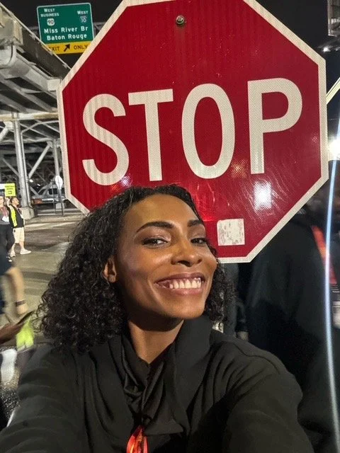 A woman smiling and taking a selfie in front of a stop sign at night. Traffic and signs visible in the background.