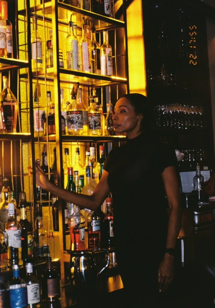 A woman with dark hair and makeup standing at a bar with a backlit display of various liquor bottles on gold shelves.