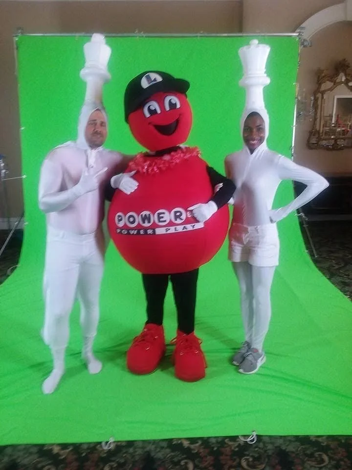 Two people dressed in futuristic white outfits with tall headpieces, standing next to a person in a round red costume with a cartoon face, a black cap, and red shoes, in front of a green screen backdrop.