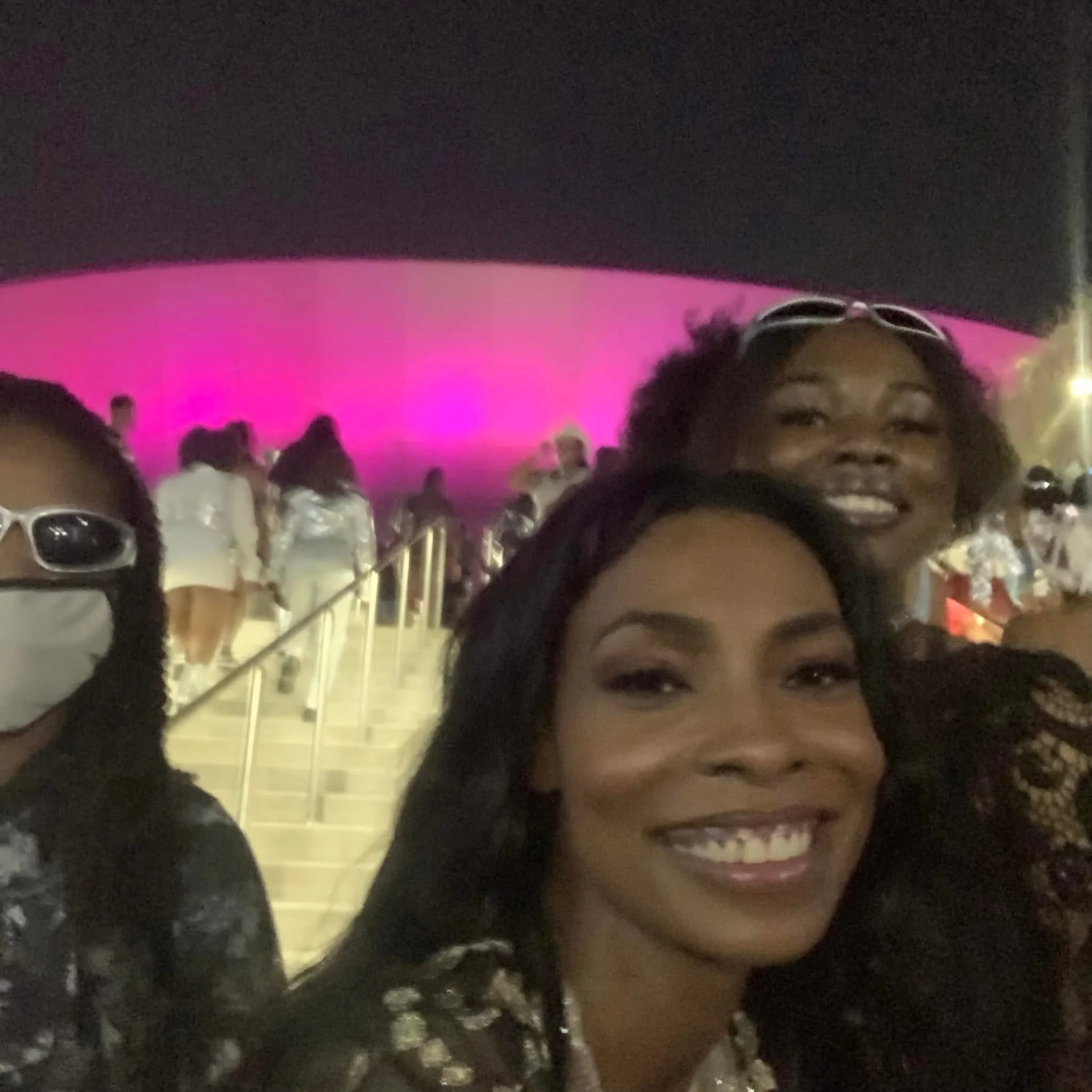 Close-up of a person's face with dark curly hair, wearing glasses, smiling in a crowd at night with bright pink and purple lighting.