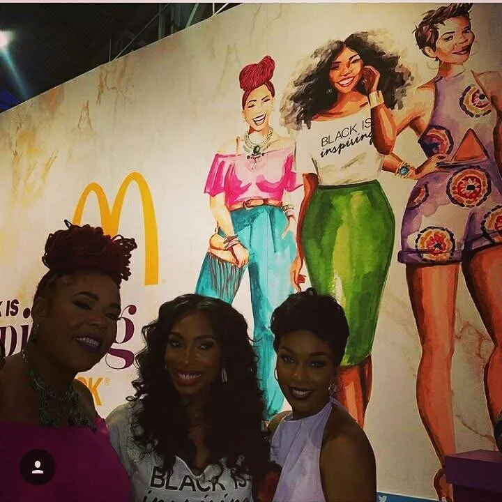 Group of three women taking a selfie in front of a colorful mural featuring diverse women in stylish outfits and a McDonald's logo.
