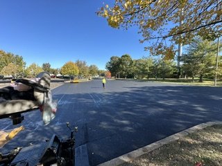 A parking lot with a worker standing in the distance, surrounded by trees with autumn foliage, under a clear blue sky.