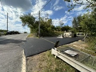 A newly paved asphalt road connecting to a dirt road, with a damaged metal fence on the side, trees, utility pole, and a small building in the background under a partly cloudy sky.