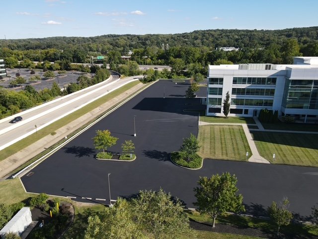 An aerial view of a modern office building with a large parking lot, surrounded by green trees and a landscaped lawn area.