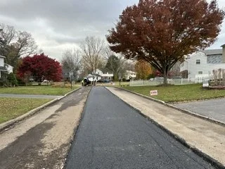 A newly paved blacktop road in a suburban neighborhood, lined with grass and trees showing fall foliage, with a cloudy sky overhead.