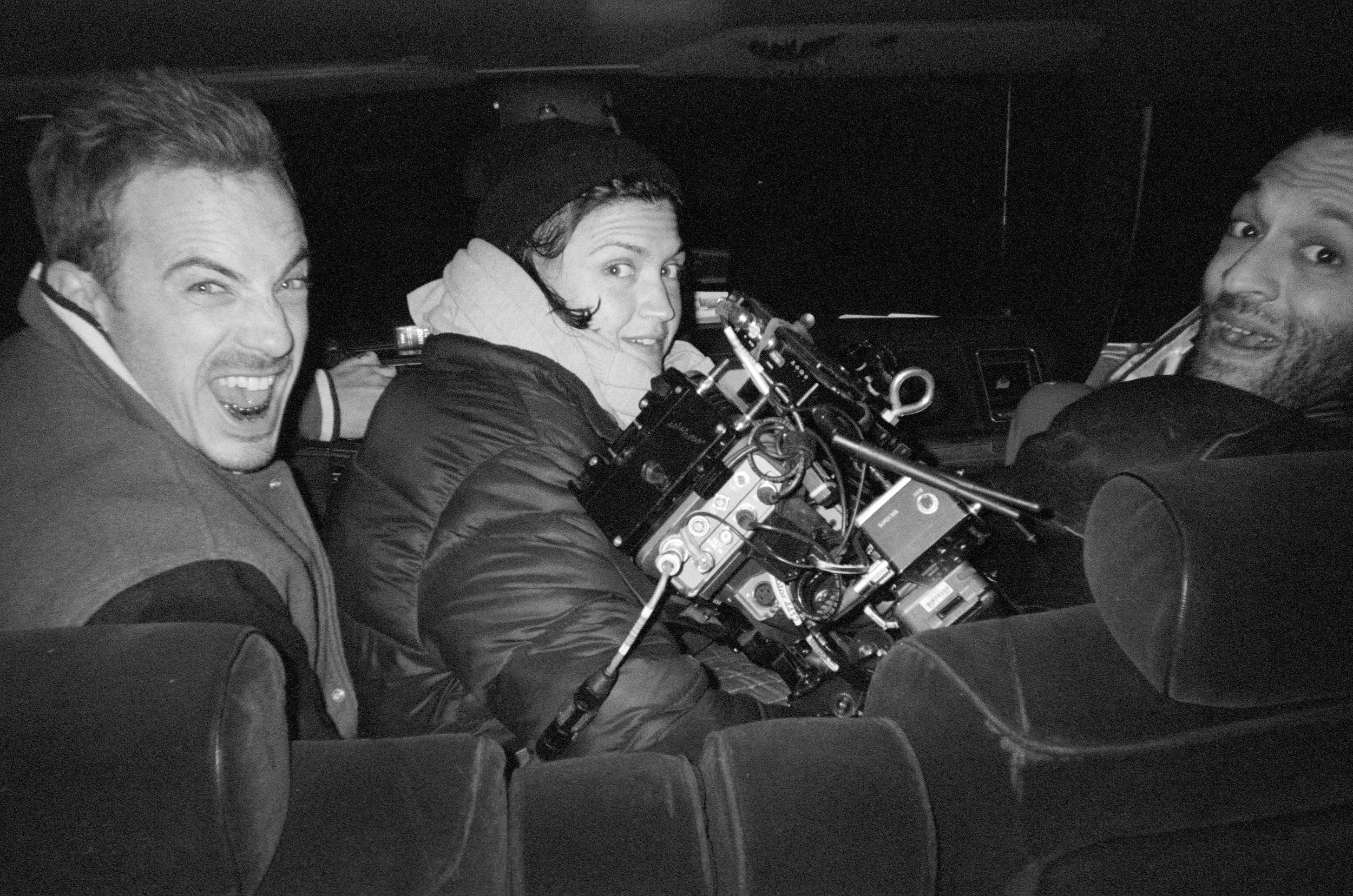 Three people sitting inside a vehicle with a professional camera setup, smiling and looking at the camera.